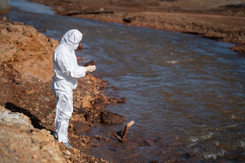 A scientist in a white protective suit takes samples of water from a polluted river.