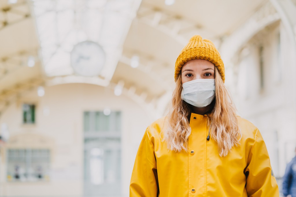Close up portrait of a young blonde in medical protective mask, yellow coat and knitted cap paralyzed with fear by news about virus cover-19. Coronavirus pandemic concept.
