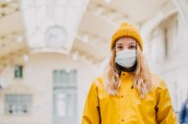 Close up portrait of a young blonde in medical protective mask, yellow coat and knitted cap paralyzed with fear by news about virus cover-19. Coronavirus pandemic concept.