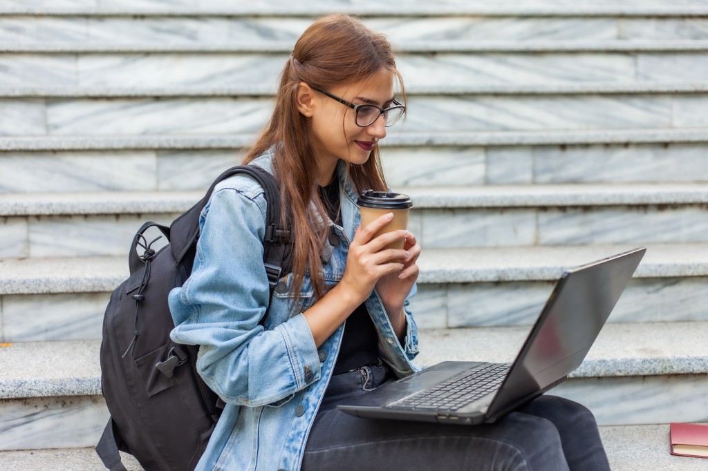 Happy modern woman student in denim jacket and backpack sitting on the stairs with laptop outdoor. Drink coffee. Watching video. Distance learning. Modern youth concept.