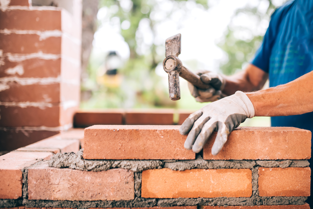 industrial worker building exterior walls, using hammer for laying bricks in cement. Detail of worker with tools